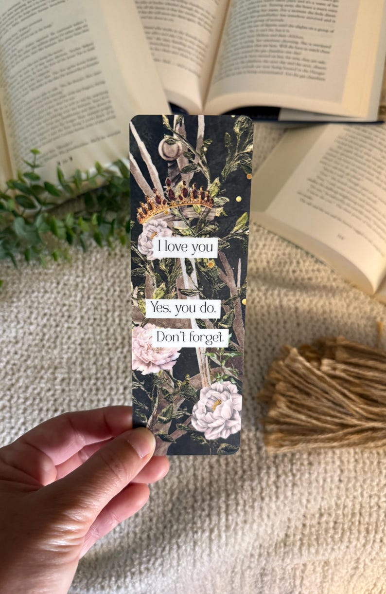 May include: A rectangular bookmark featuring a dark background with a sword, a gold crown, and pink floral accents. The text reads: I love you. Yes, you do. Don't forget. The design includes intricate botanical elements and gold speckles throughout the surface.