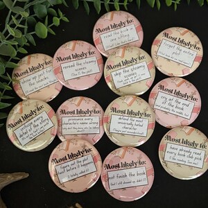 May include: A collection of eleven round pin-back badges featuring a light pink background with a pattern of books and foliage. Each badge displays the text Most likely to: followed by unique book-related phrases like read the book in one sitting.