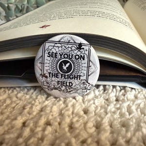 May include: A circular pin-back button featuring a black and white graphic design with a central dragon silhouette, sunburst rays, and clouds. The text reads "SEE YOU ON THE FLIGHT FIELD" in bold black capital letters within a square frame.