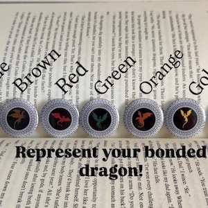 Può includere: Sette spille circolari con sagome di draghi in blu, marrone, rosso, verde, arancione, oro e nero. Ogni spilla ha un bordo bianco con linee nere radianti. Il testo sotto le spille recita, Represent your bonded dragon!