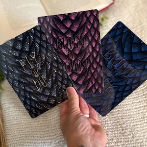 May include: Three rectangular cards featuring a dragon scale pattern in black, purple, and blue. The black card displays the text FLY OR DIE, the purple card reads BURN IT DOWN, and the blue card features the text BRING THE FIRE in gold lettering.