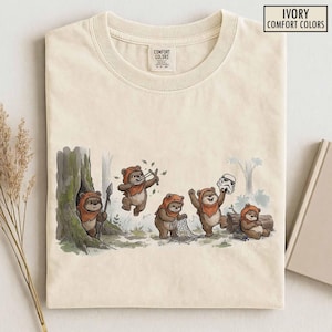 May include: An ivory crew neck t-shirt featuring a graphic illustration of five brown Ewok characters in a forest setting. One Ewok holds a spear, another uses a slingshot, one holds a net, and one holds a white stormtrooper helmet.