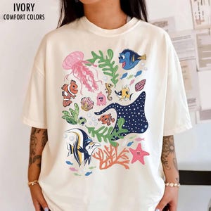 May include: An ivory short-sleeve t-shirt featuring a colorful graphic print of various sea creatures including a pink jellyfish, a blue tang, a clownfish, a sea turtle, a stingray, a starfish, and coral reefs, with the text IVORY COMFORT COLORS.