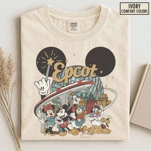May include: An ivory t-shirt featuring a graphic of the Epcot geodesic sphere, a magic wand, and Disney characters including Mickey Mouse, Minnie Mouse, Donald Duck, Goofy, Daisy Duck, and Pluto. The text reads Epcot and Ivory Comfort Colors.