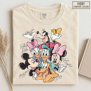 Puede incluir: Una camiseta de color marfil con un estampado gráfico de Mickey Mouse, Minnie Mouse, Donald Duck, Daisy Duck, Goofy y Pluto. El diseño incluye nombres escritos a mano para cada personaje y el texto IVORY COMFORT COLORS arriba a la derecha.