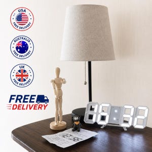 May include: A table lamp with a black base and a beige fabric shade, a wooden articulated mannequin figure, a small dark grey bear figurine, and a white digital clock displaying glowing white numbers on a dark wooden surface.