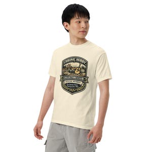 Op de afbeelding: Een crèmekleurig T-shirt met korte mouwen en een grafisch embleem op de borst. Het embleem bevat de tekst "CHRONIC HOBBY COLLECTORS CLUB OFFICIAL MEMBER" en toont diverse hobby-items zoals een gitaar, een camera en penselen in een schild.