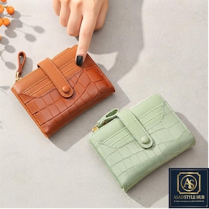 Puede incluir: Dos carteras plegables con textura de cocodrilo, una en marrón y otra en verde claro. Cada cartera incluye un cierre de botón a presión, múltiples ranuras para tarjetas y un tirador de cremallera lateral. El logo ASAD STYLE HUB es visible.