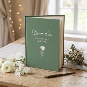 Peut inclure: Un livre d'or à couverture rigide vert sauge avec le texte blanc "Livre d'or", "Emma et Lucas" et "2 juin 2026". La couverture présente une petite illustration d'une tulipe blanche. Le livre est posé debout avec des pages blanches visibles.