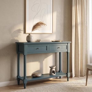 May include: A dark blue console table featuring two drawers with small round knobs and a lower shelf. The table stands on four turned legs and is topped with a light-colored vase containing dried grass, two stacked books, and a small gold tray.