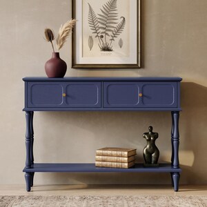 May include: A navy blue console table featuring two drawers with small gold knobs, a lower shelf, and four turned legs. On the table sits a dark red vase with dried grasses. On the lower shelf are three stacked books and a dark bronze sculpture.