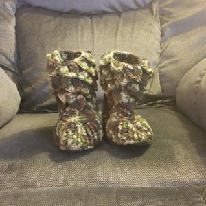 Puede incluir: Un par de zapatillas de crochet verde, marr&oacute;n y blanco con un dise&ntilde;o texturizado &uacute;nico. Las zapatillas est&aacute;n sentadas en un sof&aacute; gris.