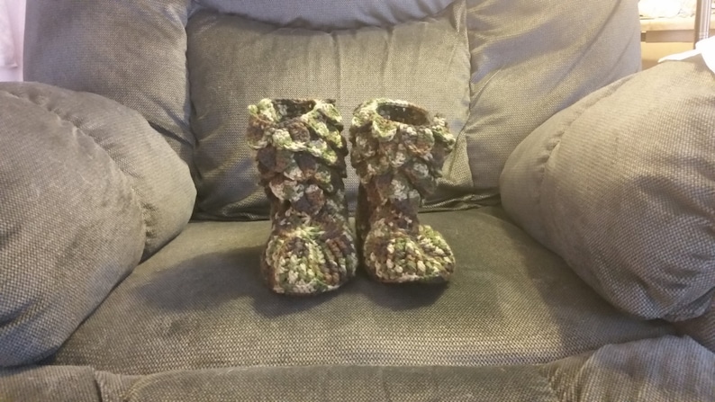 Puede incluir: Un par de zapatillas de crochet verde, marr&oacute;n y blanco con un dise&ntilde;o texturizado &uacute;nico. Las zapatillas est&aacute;n sentadas en un sof&aacute; gris.