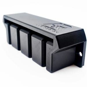 May include: A black, rectangular, wall-mounted storage rack with four vertical slots for holding items. The top surface features an embossed logo and the text "AD DESIGN". The unit has a mounting hole on the right side and a textured finish.