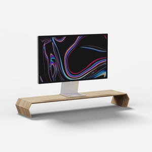 Könnte beinhalten: Ein Monitorständer aus hellem Holz mit flacher Oberseite und abgewinkelten, durchgehenden Seitenstützen. Ein Computermonitor mit dünnem silbernem Standfuß und schwarzem Bildschirm, der ein buntes, abstraktes Grafikmuster zeigt, steht darauf.