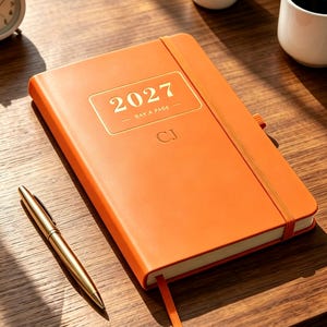 May include: An orange hardback planner with a gold elastic band closure. The cover features the text 2027 DAY A PAGE in a gold rectangular frame, with the initials CJ embossed below. A gold-toned metal pen lies diagonally next to the planner.