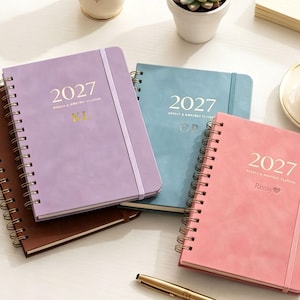 May include: A collection of four spiral-bound planners in lavender, blue, pink, and brown. Each cover features the year 2027, the text Weekly & Monthly Planner, and personalised gold or embossed lettering including KL, OP, and the name Remy with a heart icon.
