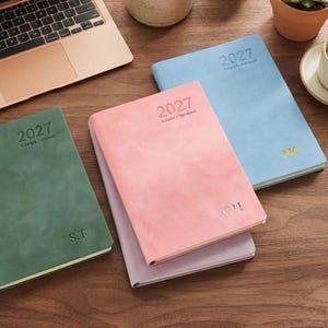 May include: Three 2027 schedule notebooks in green, pink, and light blue with a smooth matte finish. The green notebook features the initials S.T., the pink notebook features G.H., and the light blue notebook features the gold initials J.K.