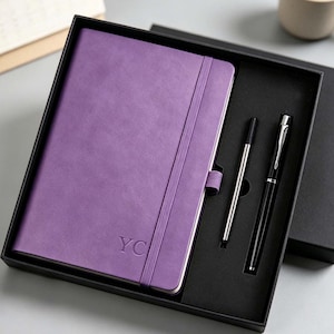 May include: A purple notebook with the embossed letters YC on the cover, featuring an elastic closure band and a pen loop. The notebook is presented in a black gift box alongside a silver-toned pen refill and a black ballpoint pen with silver trim.