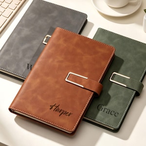 May include: Three rectangular notebooks with smooth covers in charcoal gray, tan, and dark green. Each notebook features a metallic rectangular clasp closure. The tan notebook is engraved with the name Harper, and the green one with Grace.