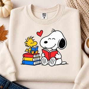 Può includere: Una felpa girocollo color crema con la grafica di un cane bianco e un uccellino giallo che leggono dei libri. Il cane tiene in mano un libro rosso, mentre l'uccellino è seduto su una pila di libri rossi, gialli e blu con un cuore rosso.