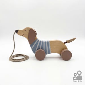 May include: A wooden pull toy shaped like a dachshund with a natural wood finish, brown ears, and a blue and white striped sweater pattern. The dog has four brown wheels and a beige pull cord attached to its nose. The text Sven & Me is visible.