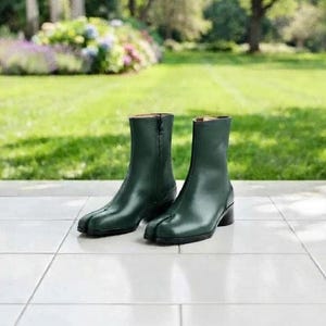 May include: A pair of dark green leather ankle boots featuring a distinct split-toe design, a side zipper closure, and a low block heel. The boots have a smooth finish and a structured silhouette that reaches mid-calf, showcasing a clean aesthetic.