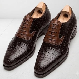 May include: A pair of dark brown oxford shoes featuring a textured crocodile skin pattern on the main body, smooth brown leather lace-up panels with brogue detailing, and light-coloured wooden shoe trees inserted into each shoe.