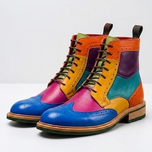 May include: A pair of lace-up ankle boots featuring a multi-colored leather design with blue, pink, yellow, orange, purple, and green panels. The boots include wingtip brogue detailing, brown laces, and a stacked wooden heel with a green sole.