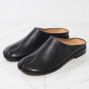 May include: A pair of black leather slip-on mules featuring a distinctive split-toe design. These shoes have a smooth, matte finish, a low heel, and a tan interior lining. The construction includes a central seam running down the top of the vamp.