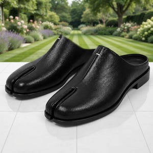 May include: A pair of black leather slip-on mules featuring a distinct split-toe design. The shoes have a smooth, textured finish, a low heel, and a single white stitch detail at the center of the vamp, creating a clean and structured appearance.