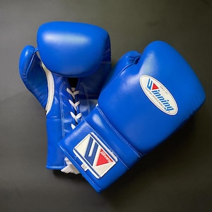 May include: A pair of bright blue boxing gloves with white lace-up closures. The gloves feature the Winning Fighting Sports World logo and a white patch on the wrist displaying the Winning logo and the text 10-OZ, indicating the weight.