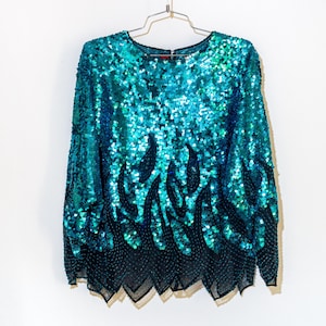 May include: A long-sleeved teal sequin top with a round neckline. The top features a flame-like design in black sequins at the bottom. The garment is hanging on a metal hanger against a white background.