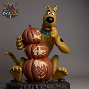 Puede incluir: Una figura marrón de Scooby-Doo con collar azul y placa, sentada detrás de una pila de tres calabazas naranjas. La calabaza superior tiene un murciélago, la del medio una furgoneta y la inferior una cara. La base es oscura y texturizada.