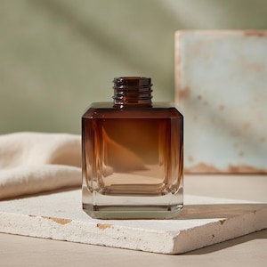 Puede incluir: Una botella de vidrio de forma cuadrada con un degradado de color que va desde un marrón ámbar intenso en la parte superior hasta una base clara y transparente. La botella tiene un cuello roscado para tapa y una base de vidrio gruesa y resistente.