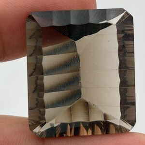 Peut inclure: Une pierre précieuse rectangulaire à facettes d'une teinte brun fumé translucide. La pierre présente une taille à degrés avec des bords nets et une surface polie qui reflète la lumière, révélant des tons bruns profonds et des zones claires.