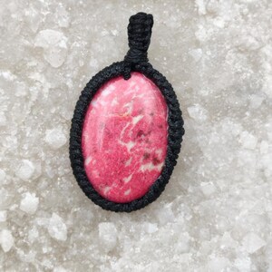 May include: An oval-shaped pendant featuring a polished pink and white mottled stone set within a black macrame cord frame. The pendant includes a matching black cord loop at the top for suspension, showcasing a textured knot pattern.
