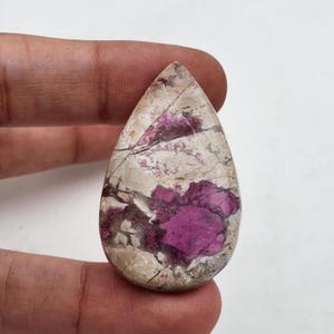 May include: A teardrop-shaped gemstone featuring a polished surface with a mix of deep magenta-pink inclusions set against a creamy off-white and light grey matrix, characterised by a network of fine, dark, natural surface fractures.