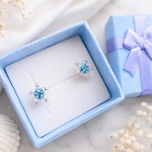 May include: A pair of silver turtle stud earrings featuring round, light blue faceted stones set in the centre of each shell. The earrings are displayed inside a square, light blue gift box with a matching ribbon bow on the lid.