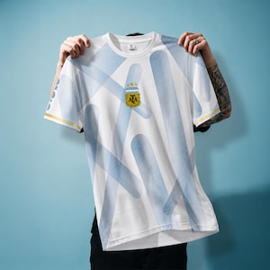 Puede incluir: Una camiseta blanca de manga corta con un patrón de líneas diagonales azul claro y el logotipo de la AFA centrado en el pecho. El logo incluye tres estrellas doradas sobre un escudo dorado con las letras AFA en negro sobre fondo azul claro.