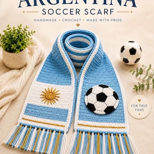 Puede incluir: Una bufanda de fútbol tejida a ganchillo a mano con los colores azul claro y blanco de la bandera argentina. Un extremo muestra un emblema de sol amarillo, mientras que el otro tiene una aplicación de balón de fútbol en blanco y negro con flecos.