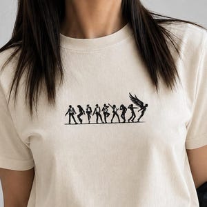 May include: A cream-colored short-sleeve t-shirt featuring a black graphic print across the chest. The design depicts a horizontal line of silhouettes showing figures in various dance poses, with the final figure on the right having large wings.