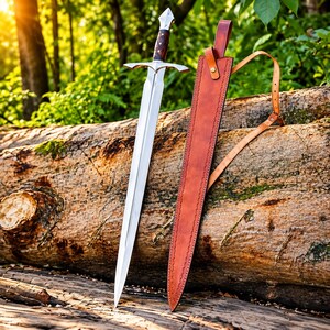 May include: A double-edged steel sword with a pointed tip, a silver crossguard, and a dark brown wooden handle with silver accents. Beside the sword is a long, tan leather scabbard featuring a matching leather strap and a brass-coloured metal buckle.
