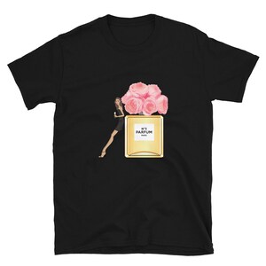 May include: Black t-shirt featuring a graphic of a perfume bottle with pink roses and a woman in a black dress. The perfume bottle is gold with the text "N&deg;5 PARFUM PARIS".
