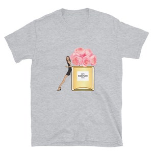 May include: A light gray t-shirt featuring a graphic of a woman in a black dress, a perfume bottle with pink roses, and the text "N&deg;5 PARFUM PARIS". The design is centered on the shirt.