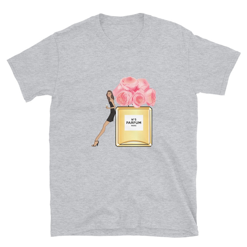 May include: A light gray t-shirt featuring a graphic of a woman in a black dress, a perfume bottle with pink roses, and the text "N&deg;5 PARFUM PARIS". The design is centered on the shirt.