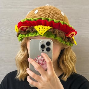 May include: A crochet bucket hat designed to resemble a hamburger, featuring a tan bun top with white sesame seed accents, layered with ruffled red tomato, brown meat patty, yellow cheese, and green lettuce textures around the brim.