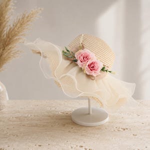 May include: A wide-brimmed straw hat featuring a natural tan woven texture, adorned with two soft pink fabric roses and green leaves. The brim is layered with a voluminous, ruffled cream-coloured organza trim that creates a delicate, wavy edge.