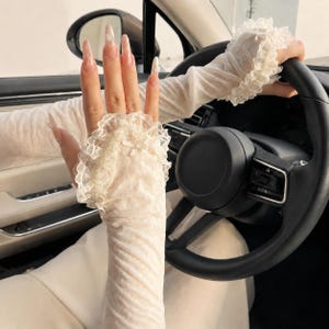 Peut inclure: Une paire de manchettes sans doigts, transparentes et blanc cassé, confectionnées dans un tissu léger et texturé. Les manches présentent un bord en dentelle volantée au poignet et au bras, agrémenté d'un petit nœud délicat sur le dos de la main.