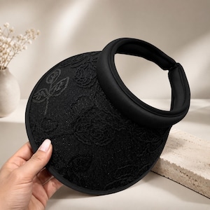 May include: A black sun visor featuring an intricate embroidered floral pattern across the wide brim. The visor has a smooth, padded headband and a structured, rounded shape, providing a clear view of the detailed stitching on the fabric.